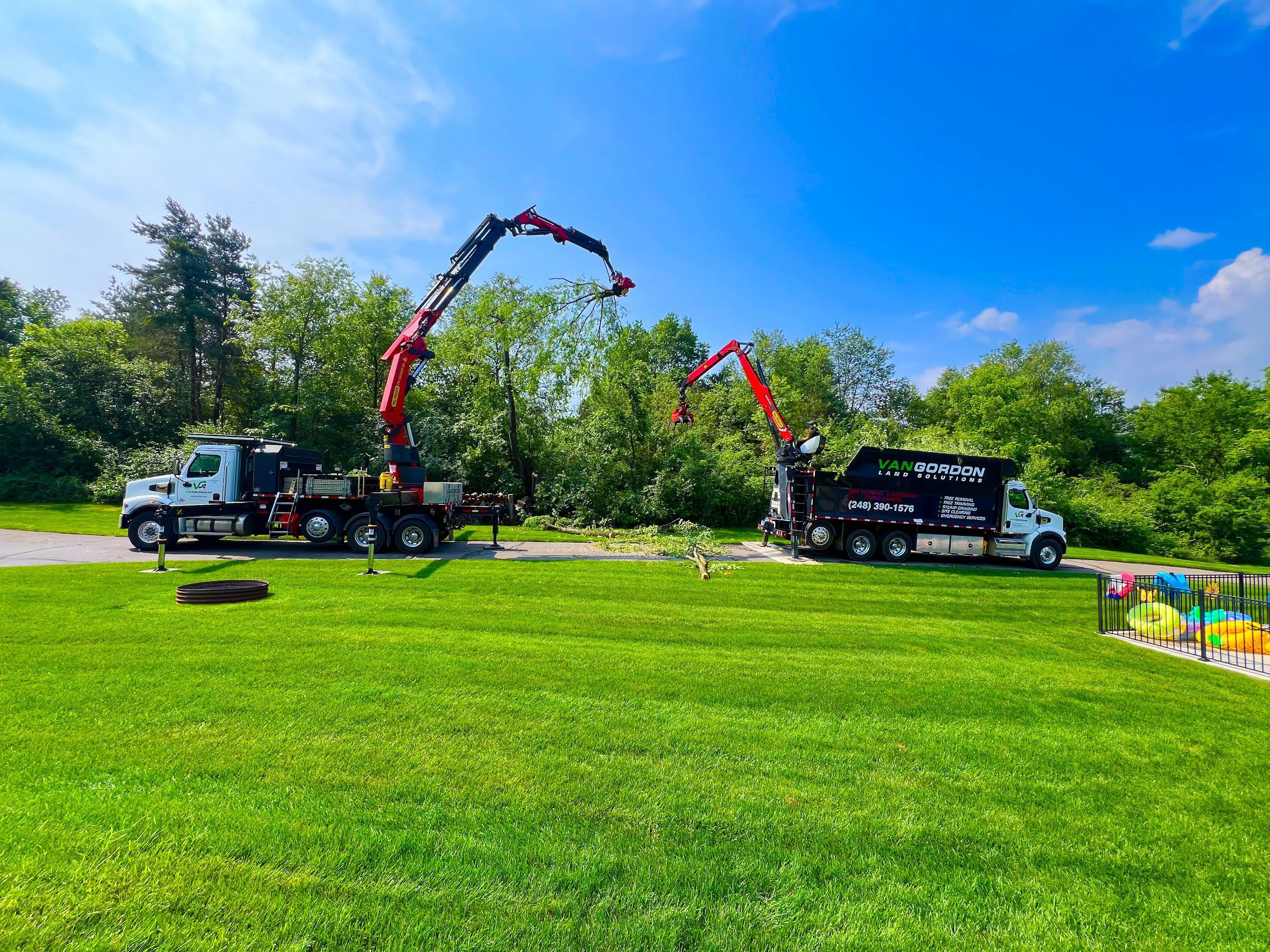 Crane-assisted tree removal trucks lift large branches safely away from homes in a clear, green yard.